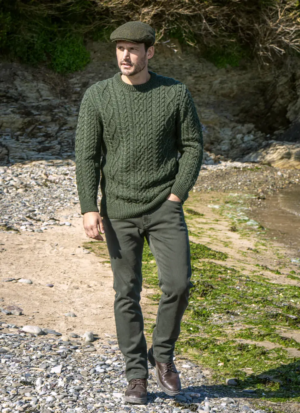 Man Walking On Beach in Paddy Cap Wearing Oisin Aran Crew Neck Sweater in Army Green